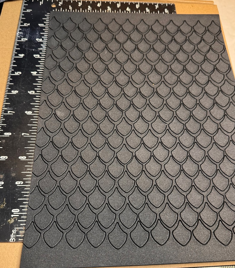 Op de afbeelding: Zwarte, getextureerde plaat met een herhalend patroon van verhoogde, overlappende schubben. De schubben zijn in een verspringend patroon gerangschikt, waardoor een subtiel 3D-effect ontstaat.
