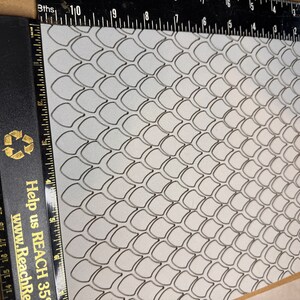 Op de afbeelding: Een wit vel papier met een zwart schubbenpatroon erop gedrukt. Het papier ligt op een bruin oppervlak en een zwarte liniaal is langs de linkerkant van het papier geplaatst.