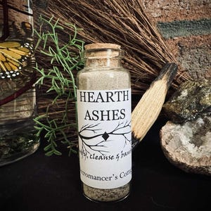 May include: A glass jar labeled "HEARTH ASHES" with a cork top, filled with gray ash. The label includes the text "purify, cleanse & banish" and "romancer's Cottage". A bundle of dried herbs and a piece of wood are next to the jar.