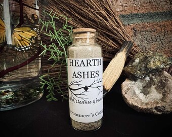 May include: A glass jar labeled "HEARTH ASHES" with a cork top, filled with gray ash. The label includes the text "purify, cleanse & banish" and "romancer's Cottage". A bundle of dried herbs and a piece of wood are next to the jar.
