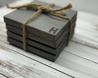 Puede incluir: Conjunto de cuatro posavasos de madera gris con una sola inicial "H" quemada en el posavasos superior. Los posavasos están atados con cuerda.