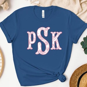 May include: A royal blue t-shirt with a pink floral monogram that reads "PSK".
