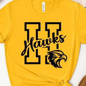 May include: Mustard yellow t-shirt with a black graphic. The graphic features the letter "H" twice, the word "Hawks" in a script font, and a stylized eagle head. The shirt is made of a soft material.