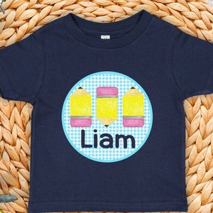 May include: Navy blue toddler t-shirt featuring a circular design with three yellow pencils with pink erasers on a blue and white gingham background. The name "Liam" is printed in white below the pencils.