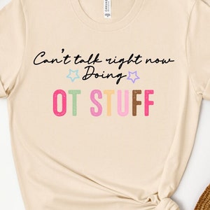 May include: A beige t-shirt with the text "Can't talk right now Doing OT STUFF" in black and colorful lettering.