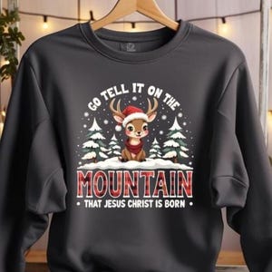 May include: Dark gray sweatshirt with a Christmas graphic. The graphic features a cartoon deer wearing a Santa hat and a scarf, with the text "GO TELL IT ON THE MOUNTAIN THAT JESUS CHRIST IS BORN" in white and red. The sweatshirt is hanging on a wooden hanger.