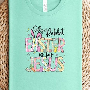 May include: A light teal t-shirt with the text "Silly Rabbit Easter is for Jesus." The text features a bunny, carrots, and Easter eggs. The word "Easter" is in a pink and white plaid pattern.