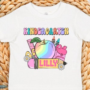 May include: White t-shirt with a rainbow apple, pencil, ruler, and backpack design. The text "KINDERGARTEN" is above the design and the name "LILLY" is on the pencil.