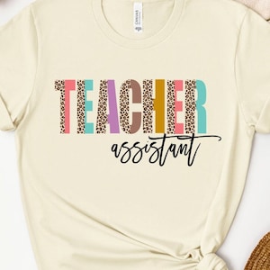 May include: A light beige t-shirt with the text "TEACHER assistant" in black lettering. The word "TEACHER" is in colorful letters with a leopard print pattern.