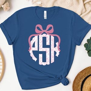 May include: A royal blue t-shirt with a pink monogram featuring the letters "ASH" in a circle with a pink bow on top. The monogram is surrounded by a white scalloped border and a floral pattern.