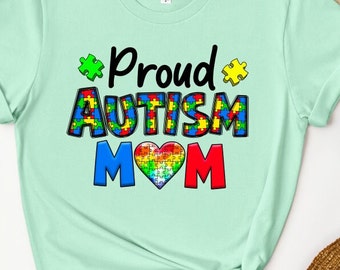 May include: A light green t-shirt with the text "Proud Autism Mom" in black. The word "Autism" is made up of colorful puzzle pieces and a rainbow puzzle piece heart is between the "M"s.
