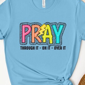 May include: Light blue t-shirt with a colorful "PRAY" graphic with black polka dots. The text "THROUGH IT - ON IT - OVER IT" is printed below the graphic.