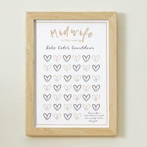 May include: A framed print with the words "Midwife in the making" and "Baby Catch Countdown" in gold script. The print features a grid of heart shapes, some filled in, with a reminder at the bottom. The frame is made of light-colored wood.