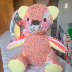May include: A pink and yellow patchwork teddy bear with floral fabric accents. The bear has a crocheted body and a sweet expression.