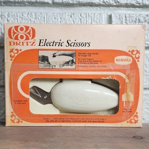 May include: White electric scissors with a silver blade, packaged in a box with orange and white accents. The box features the brand name "DRITZ" and the text "Electric Scissors". The scissors are labelled "DRITZ Electric Scissors" and have a cord that plugs into an outlet. The box also includes the text "Complete with 8 foot cord" and "Quick... cuts cutting time - Helps eliminate hand and wrist fatigue. Use with left or right hand - Cuts both sheer and heavy fabrics!"