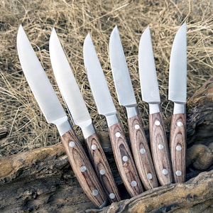 May include: Set of six silver steak knives with wooden handles. The handles have a dark brown wood grain pattern.