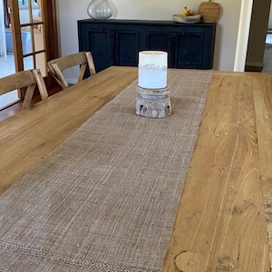 May include: A dining table with a brown woven table runner. A white and blue candle sits on a wooden stand. The table is made of light-coloured wood, and the runner has a fringed edge. The table is in a room with a dark blue cabinet.