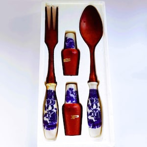 Può includere: Un set di posate da portata in scatola, tra cui una forchetta e un cucchiaio con manici in legno scuro e basi in porcellana bianca decorate con motivi floreali blu. Sono incluse anche due piccole bottiglie rosse con dettagli dorati.