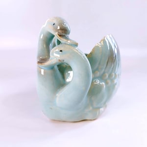 May include: A light blue ceramic planter shaped like two swans with their necks curved towards each other. The glossy finish features subtle brown shading on the beaks and wings, with moulded feather details on the sides and a hollow centre.