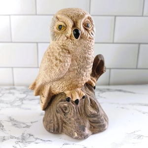May include: A detailed ceramic owl figurine perched on a tree stump. The owl has tan and brown coloring with yellow eyes and a black beak. The tree stump is brown with a textured, wood-like appearance. A decorative item.