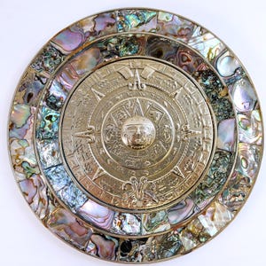 May include: A silver-toned Aztec calendar with intricate carvings, centered on a round plate. The outer ring is inlaid with iridescent abalone shell pieces in shades of blue, green, and purple. The design features a sun face and detailed symbols.