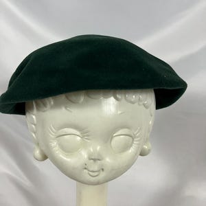 May include: A dark green beret. The hat is made of a soft, felt-like material and has a classic, round shape. The beret is displayed on a vintage mannequin head.