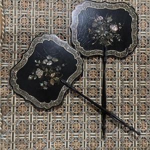 May include: Two black decorative hand fans with floral designs. The fans have a scalloped edge with a silver trim. The floral designs are made of mother of pearl. The fans are on a woven background.