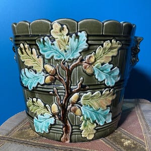 May include: A green ceramic planter with a detailed oak leaf and acorn design. The planter features a textured surface and a scalloped rim. The leaves are in shades of blue, green, and yellow, with brown acorns and branches. Decorative handles are on the sides.