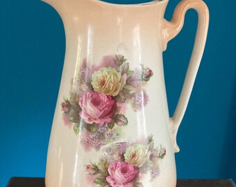 May include: A vintage cream-coloured pitcher with a floral design featuring pink and yellow roses. The pitcher has a wide mouth and a handle.