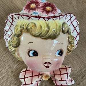 May include: A vintage ceramic wall planter shaped like a girl's head. The planter has a pink and white checked bow, a pink and white checked hat with pink flowers, and a painted face with blue eyes.