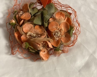 Puede incluir: Tocado floral vintage con flores de tela color melocotón, hojas verdes y una red naranja quemado. Un lazo de terciopelo naranja quemado acentúa la parte superior. El tocado es probablemente para una ocasión especial.