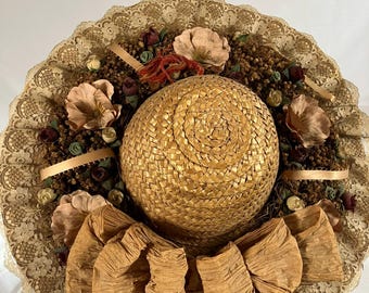 May include: A straw hat featuring a wide brim edged with intricate beige lace. The crown is surrounded by a cluster of dried brown foliage, small fabric flowers in muted tones, and a large, textured tan ribbon bow at the front base.