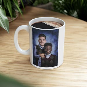 May include: White ceramic coffee mug featuring a portrait of two smiling people wearing argyle vests over collared shirts. The mug is filled with dark coffee and has a white handle. The background is a gradient of blue.