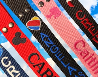 May include: Five fabric wristlet keychains in red, black, pink, and navy blue. Each strap features silver metal snaps and embroidered designs including a Mickey Mouse silhouette, a heart, a doll profile, and names like Angela and Caitlin.