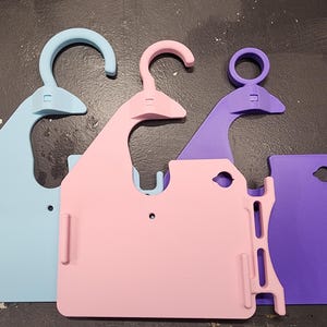 May include: Three colorful, plastic hangers in light blue, pink, and purple. Each hanger has a hook and a rectangular base with a slot and a small hole. The hangers are designed for holding items.