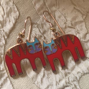 May include: A pair of cat-shaped earrings. Each earring features a red cat with a blue and purple face, and a copper-colored outline. The earrings have a hook closure and are set against a textured background.