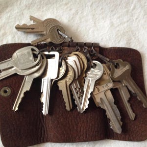 May include: A collection of keys attached to a brown leather key holder. The keys are various sizes and shapes, with some bearing manufacturer markings. The key holder has a snap closure and a worn appearance.