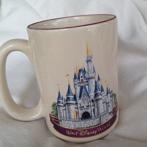 May include: A white ceramic mug with a red rim features a colorful illustration of Cinderella's Castle at Walt Disney World. The castle is surrounded by a purple banner with the text "Walt Disney World".