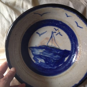 May include: A white ceramic bowl with a blue rim and a hand-painted design of a sailboat on the water with seagulls flying overhead.