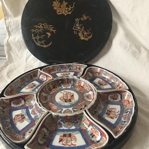 May include: A black, round, lidded serving dish with a set of eight decorative plates. The lid features gold floral designs. The plates are white, blue, and red with intricate patterns and a central design. The set is ideal for serving appetizers or small dishes.