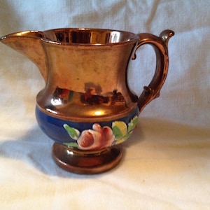 May include: A copper-colored ceramic pitcher with a handle and spout. The base features a blue band with a floral design of pink roses and green leaves. The pitcher has a shiny, reflective surface.