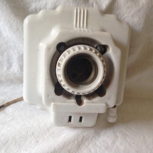 May include: White porcelain light fixture with a single socket and two slots for wiring. The fixture has a decorative design with three vertical lines on the top.