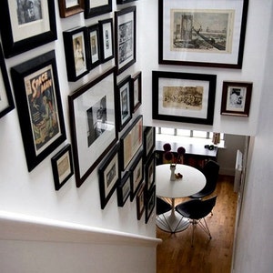 May include: A white wall with a gallery wall of black framed pictures and prints. The pictures are of various sizes and shapes and are arranged in a random pattern. The wall is in a home and there is a white table and chairs in the background.