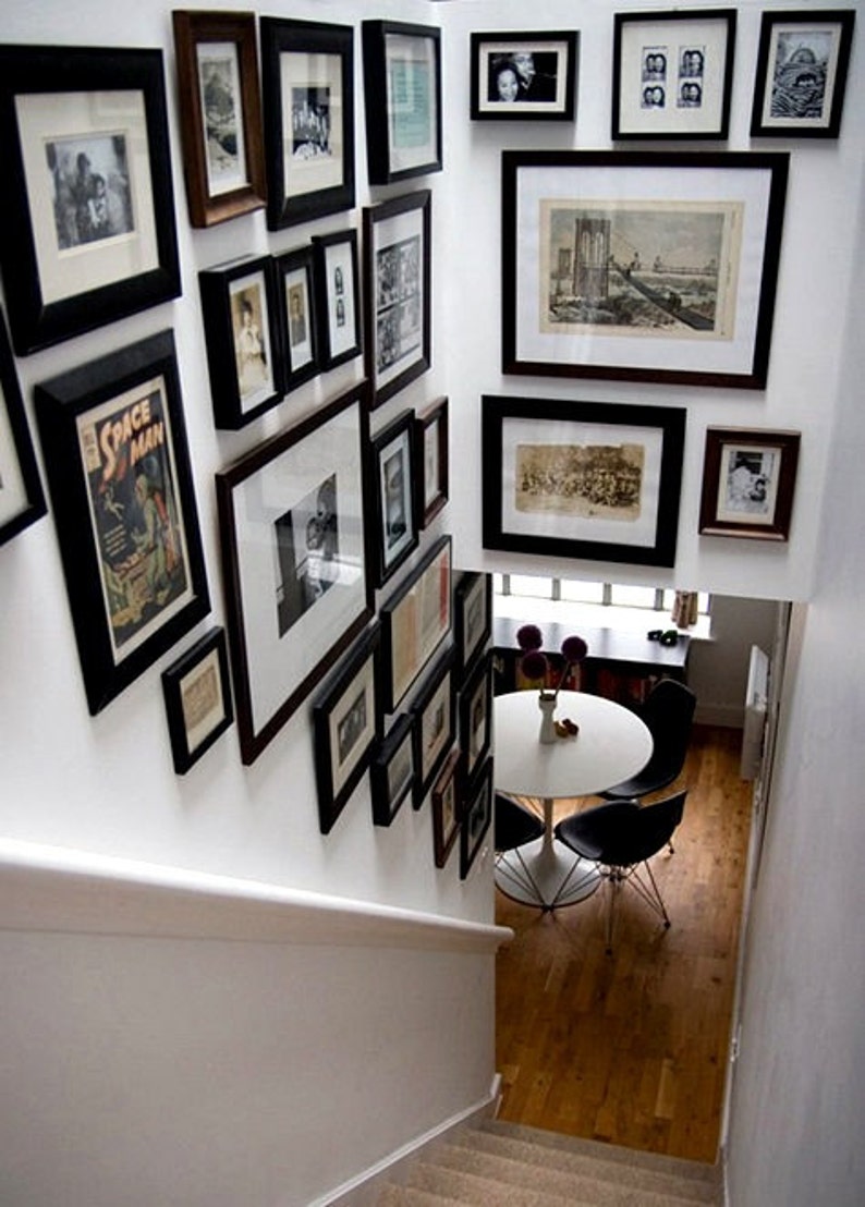 May include: A white wall with a gallery wall of black framed pictures and prints. The pictures are of various sizes and shapes and are arranged in a random pattern. The wall is in a home and there is a white table and chairs in the background.