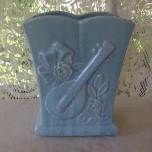 May include: A light blue ceramic vase with a raised design of a guitar, a bow, and roses. The vase has a speckled glaze.