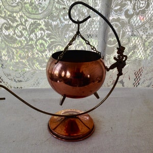 May include: A copper hanging planter with a whimsical figure on the side. The planter is suspended from a chain and sits on a copper stand with a curved handle.
