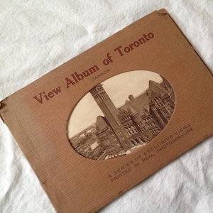 May include: A vintage View Album of Toronto, Canada, with a sepia-toned image of a building visible through an oval cutout. The album cover is a faded brown color, with text in a serif font.