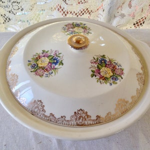 May include: A white ceramic serving dish with a lid, featuring a floral design with gold accents. The dish has two handles and a gold knob on the lid.