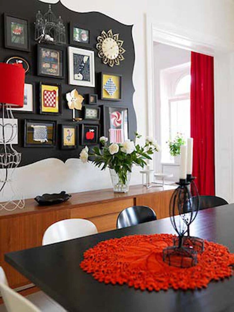 May include: A black dining table with a red crocheted table runner and a black metal candle holder with white candles. The table is set with white chairs and a wooden sideboard with a black and white patterned wall behind it.