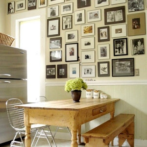 May include: A light wood dining table with a single drawer and a matching bench. The table has turned legs and is set against a wall covered in framed black and white photos.